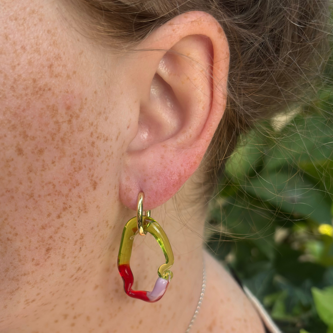 A close-up of a person's ear wearing a chunky, open-ended hoop earring made of translucent lime green, red, and lavender acrylic with a gold-tone top loop.