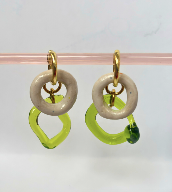 Pair of earrings with gold loops, beige circular middle pieces, and green open rings on a pink stand.