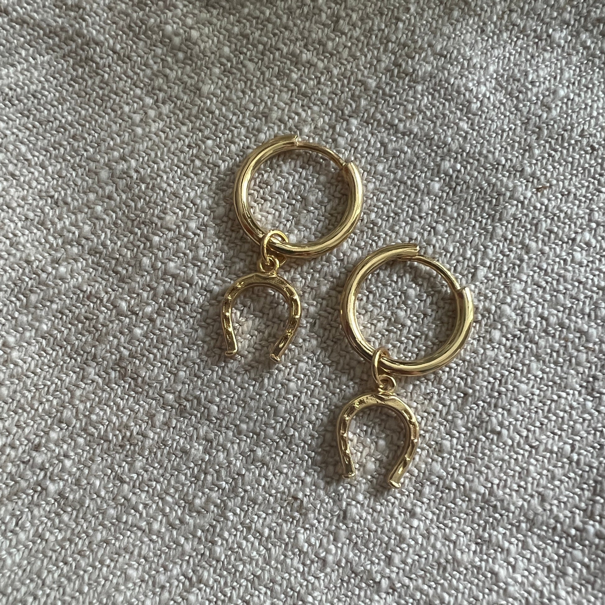Gold hoop earrings with a horse shoe charm. The horse shoe faces downwards to represent good luck. The photo is on a plain cream linen rough texture. 